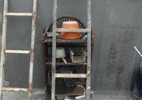 Random small industrial tools equipment on rusty shelf. Orange brown upside down bucket with multiple small trinkets and old rusty steel ladder on gray concrete floor and walls. photo