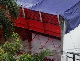 West Jakarta, Indonesia - February 18th, 2023 - House store with white walls and net red gate and blue terpal roof isolated with green tree decoration. photo