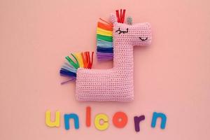 Crochet amigurumi handmade stuffed soft pink unicorn toy with rainbow mane and word on pink background. Handwork hobby. Craft diy newborn pregnancy concept. Knitted doll for little baby. Flat lay photo