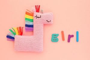 Crochet amigurumi handmade stuffed soft pink unicorn toy with rainbow mane and word Girl on pink background. Handwork hobby. Craft diy newborn pregnancy concept. Knitted doll for little baby. Flatlay photo