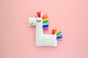 Crochet amigurumi handmade stuffed soft white unicorn toy with rainbow mane on pink background. Handwork, hobby. Craft diy newborn pregnancy concept. Knitted doll for little baby. Close up flat lay photo