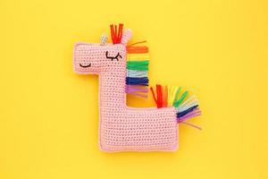 Crochet amigurumi handmade stuffed soft pink unicorn toy with rainbow mane on yellow background. Handwork, hobby. Craft diy newborn pregnancy concept. Knitted doll for little baby. Close up flat lay photo