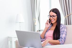 Happy woman sitting on sofa with laptop and talking on phone at home. Young successful businesswoman working from home while talking at phone. College student studying on laptop and using phone. photo