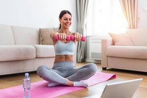 Young woman exercise with dumbbells at home and watching tutorial lesson on laptop computer. happy young female doing arm workout using dumbbells and looking at tutorial on her laptop photo