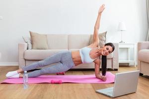 Portrait of young attractive woman doing exercises while watching online lessons on her laptop. Healthy lifestyle and sports concept. Shot of fitness woman sitting on exercise mat photo