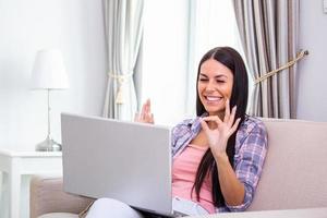 Attractive happy young woman sitting on the sofa using laptop computer, having video chat, waving, talks with her friend about coronavirus and stay home during online video call. Working home photo