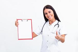 Portrait of a young female doctor in a lab coat poses with a clipboard in hand showing thumbs up. Isolated on white. photo
