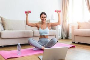 Young woman exercise with dumbbells at home and watching tutorial lesson on laptop computer. happy young female doing arm workout using dumbbells and looking at tutorial on her laptop photo