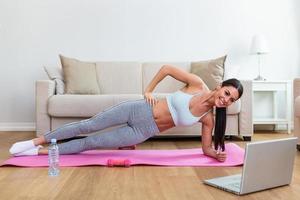 Girl training at home, doing workout and watching videos on laptop, training in living room. Beautiful woman is doing side plank exercise. She is watching video on the internet and repeating the tasks photo