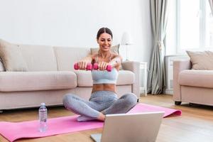 Young woman exercise with dumbbells at home and watching tutorial lesson on laptop computer. happy young female doing arm workout using dumbbells and looking at tutorial on her laptop photo