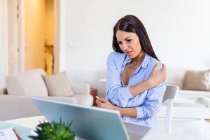 Portrait of young stressed woman sitting at home office desk in front of laptop, touching aching shoulder with pained expression, suffering from shoulder after working on laptop photo