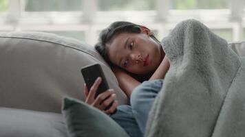 imágenes de infeliz triste asiático mujer en capa sufrimiento desde fiebre y gripe en sofá mientras utilizando móvil teléfono en el vivo habitación. salud cuidado y médico conceptos. video