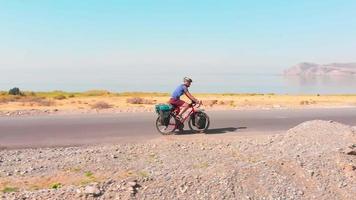 Aerial side tracking view young fit caucasian male cyclist cycle fully loaded bicycle tour bike by scenic lake background in sunny summer day. Active holidays and travel in Armenia video