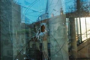 A hole in the window glass with a bullet during a military shooting. Cracks propagate around the hole. Dirty window frame. View of the street from the inside. photo