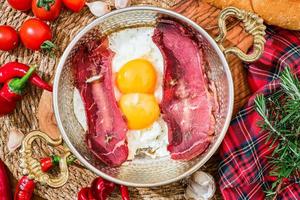 Sliced Turkish bacon pastrima for breakfast, fried beef with eggs peppers and vegetables in a traditional copper Turkish pan, top view, local cuisine. photo