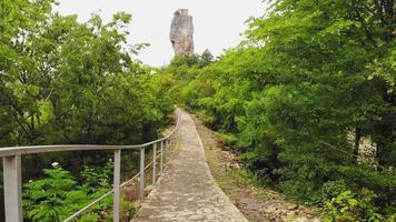 Stone walkway in forest outdoors on hilly terrain with background of Katskhi pillar with iconic church high up on top. Georgian meteora monastery concept. video