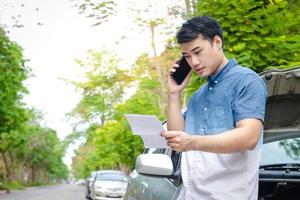 An Asian man has crashed into a car accident. Park your car on the road, hold paperwork, and call the car insurance company. Concept of insurance. Copy space photo