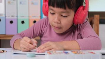 alegre pequeño niña en auriculares canto y dibujo con de colores lapices en papel sentado a mesa en su habitación a hogar. creatividad y desarrollo de multa motor habilidades. video