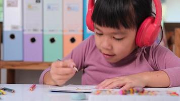 Cheerful little girl in headphones singing and drawing with colored pencils on paper sitting at table in her room at home. Creativity and development of fine motor skills. video