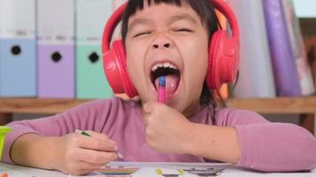 de bonne humeur peu fille dans écouteurs en chantant et dessin avec coloré des crayons sur papier séance à table dans sa pièce à maison. la créativité et développement de bien moteur compétences. video