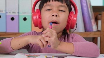 alegre pequeño niña en auriculares canto y dibujo con de colores lapices en papel sentado a mesa en su habitación a hogar. creatividad y desarrollo de multa motor habilidades. video