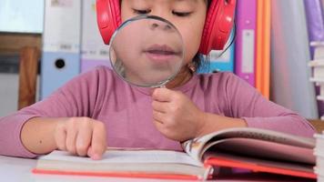 alegre pequeño niña en auriculares canto mientras leyendo un libro se sienta a el mesa en su habitación a hogar. aficiones para niños a hogar video