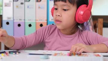 alegre pequeño niña en auriculares canto y dibujo con de colores lapices en papel sentado a mesa en su habitación a hogar. creatividad y desarrollo de multa motor habilidades. video