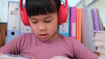 alegre pequeño niña en auriculares leyendo un libro se sienta a el mesa en su habitación a hogar. tecnología y aficiones para niños video