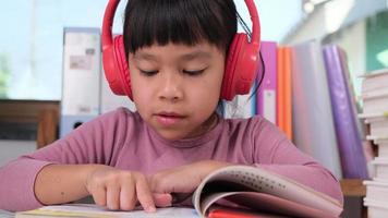 alegre pequeno menina dentro fones de ouvido lendo uma livro senta às a mesa dentro dela quarto às lar. tecnologia e hobbies para crianças video