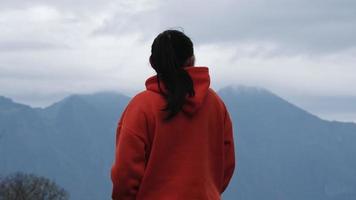 posterior ver de un joven mujer en pie en el parte superior de un calma montaña y mirando a el Mañana nubes mujer vistiendo un suéter disfrutando el belleza de naturaleza mirando a el montaña en invierno. video