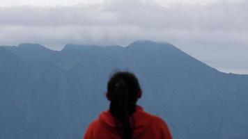 posterior ver de un joven mujer en pie en el parte superior de un calma montaña y mirando a el Mañana nubes mujer vistiendo un suéter disfrutando el belleza de naturaleza mirando a el montaña en invierno. video