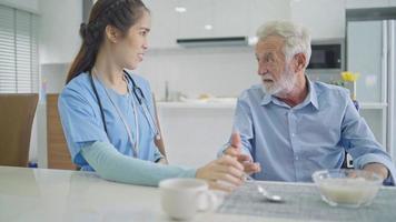 Patient senior man talking with female caregiver during eating breakfast. Female nurse talking care of male elderly at nursing home video