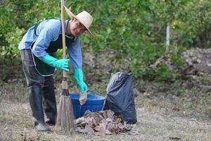 Asian male  gardener holds broom , basket, black garbage bag to clean dry leaves in garden.  Conceprt, get rid of dry leaves to make compost or make fire barrier in autumn. photo