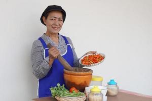 feliz anciana asiática está cocinando, usa gorro de chef y delantal, sostiene mortero, mortero y plato de chiles. concepto, cocinar para la familia. estilo de vida de la cocina tailandesa. actividad de ancianos. foto