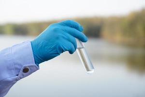 Closeup researcher hands wears blue gloves holds test glass tube that contain sample water from the lake. Concept, explore, inspect quality of water from natural source. photo