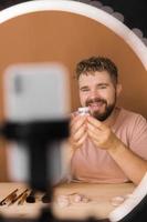 Portrait of happy man beauty blogger sitting in room at table and speaking recommending foundation or decorative cosmetic looking at camera. Blogging and vlog concept photo