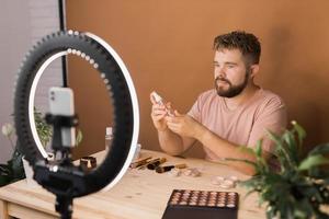 Portrait of happy man beauty blogger sitting in room at table and speaking recommending foundation or decorative cosmetic looking at camera. Blogging and vlog concept photo