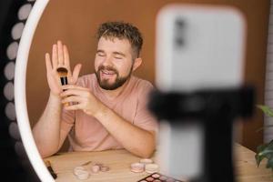 Portrait of happy man beauty blogger sitting in room at table and speaking recommending foundation or decorative cosmetic looking at camera. Blogging and vlog concept photo