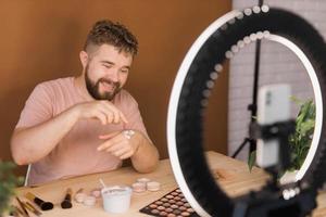 Portrait of happy man beauty blogger sitting in room at table and speaking recommending foundation or decorative cosmetic looking at camera. Blogging and vlog concept photo