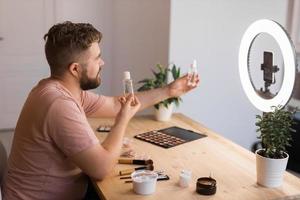 Portrait of happy man beauty blogger sitting in room at table and speaking recommending foundation or decorative cosmetic looking at camera. Blogging and vlog concept photo