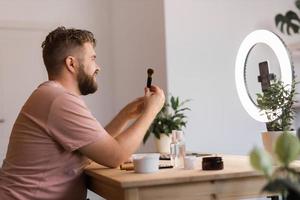 Portrait of happy man beauty blogger sitting in room at table and speaking recommending foundation or decorative cosmetic looking at camera. Blogging and vlog concept photo