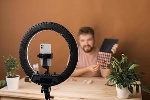 Portrait of happy man beauty blogger sitting in room at table and speaking recommending foundation or decorative cosmetic looking at camera. Blogging and vlog concept photo