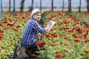 Asian gardener is taking note using clip board on the growth and health of red zinnia plant while working in his rural field farm for medicinal herb and cut flower concept photo