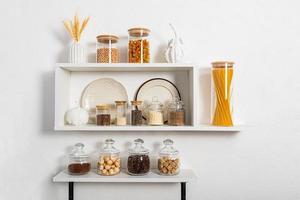 a set of glass and ceramic dishes, glass filled jars with bulk products on white kitchen shelves on textured wall. compact storage. Stylish design. photo