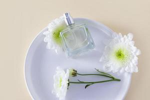 a transparent bottle of perfume or toilet water lies on a white chrysanthemum flower and a ceramic plate. top view. beige background. photo