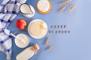 different types of dairy products in bowls, bottles and jugs on a blue background. top view. flat lay. wooden letters with the text shavuot. photo