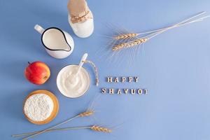 festivo antecedentes para el judío fiesta de shavuot. lechería productos, manzana, orejas de maíz en un azul antecedentes con un congratulatorio texto. parte superior vista. foto