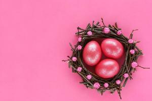 Three 3 Easter eggs colored pink shiny color in nest made of tree branches decorated with pom poms on pink paper background. Glitter paint for eggs. Greeting card for easter holiday. Place for text. photo