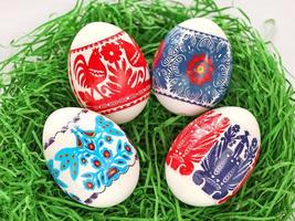 Four Colourful Easter Eggs with floral, birds, people folk art pattern printed on stickers. Eggs in green paper grass nest. Polish traditional paper cut from Kurpie region. photo