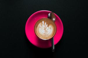 caliente latté en rosado taza con floral modelo en espuma parte superior ver en un oscuro antecedentes foto